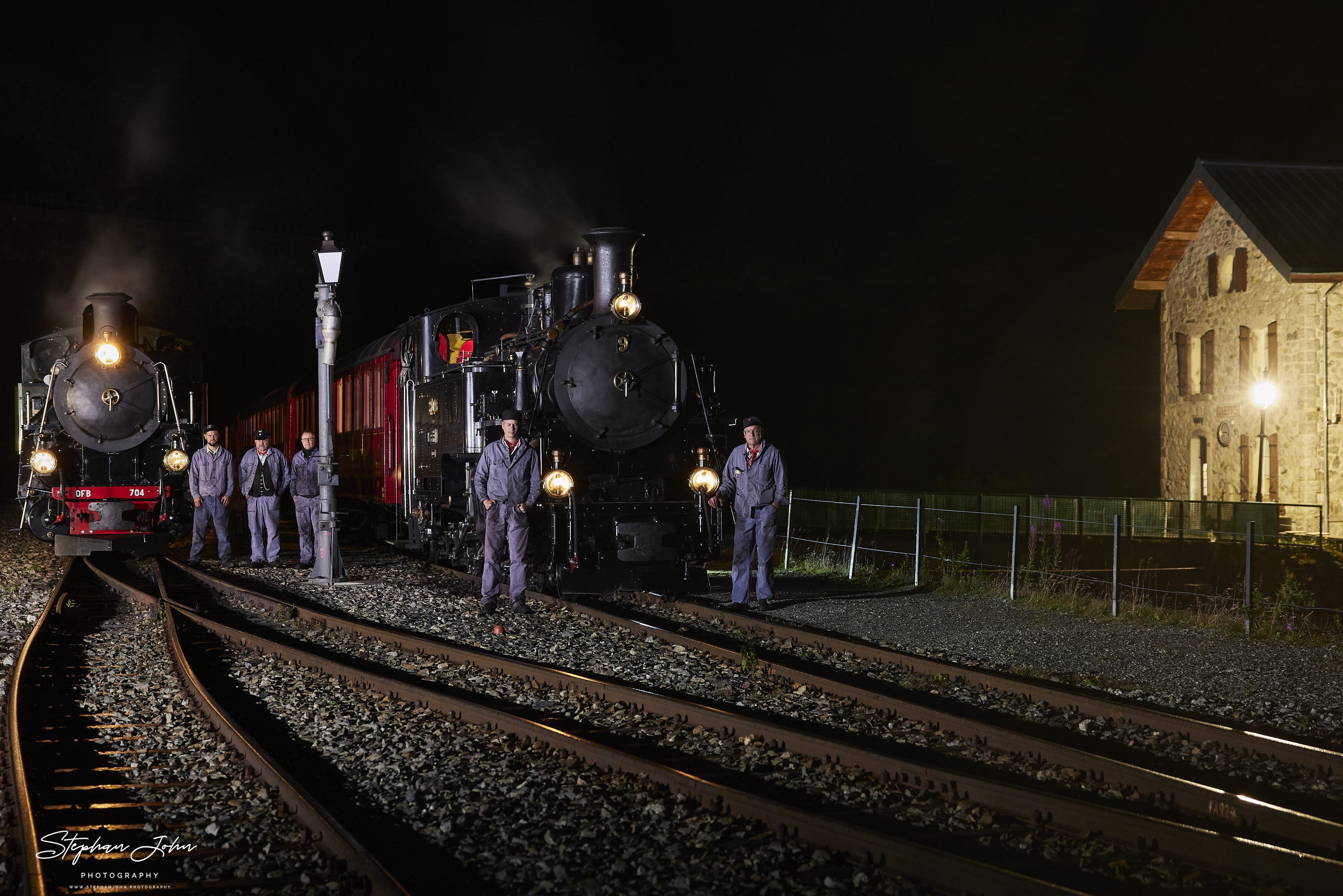 Bahnhog Gletsch am Abend. Lok 9 mit einem Personenzug und Lok 704 mit einem Güterzug stehen abfahrtbereit in Richtung Furka. Die Lokmannschaften posieren vor den Zügen.