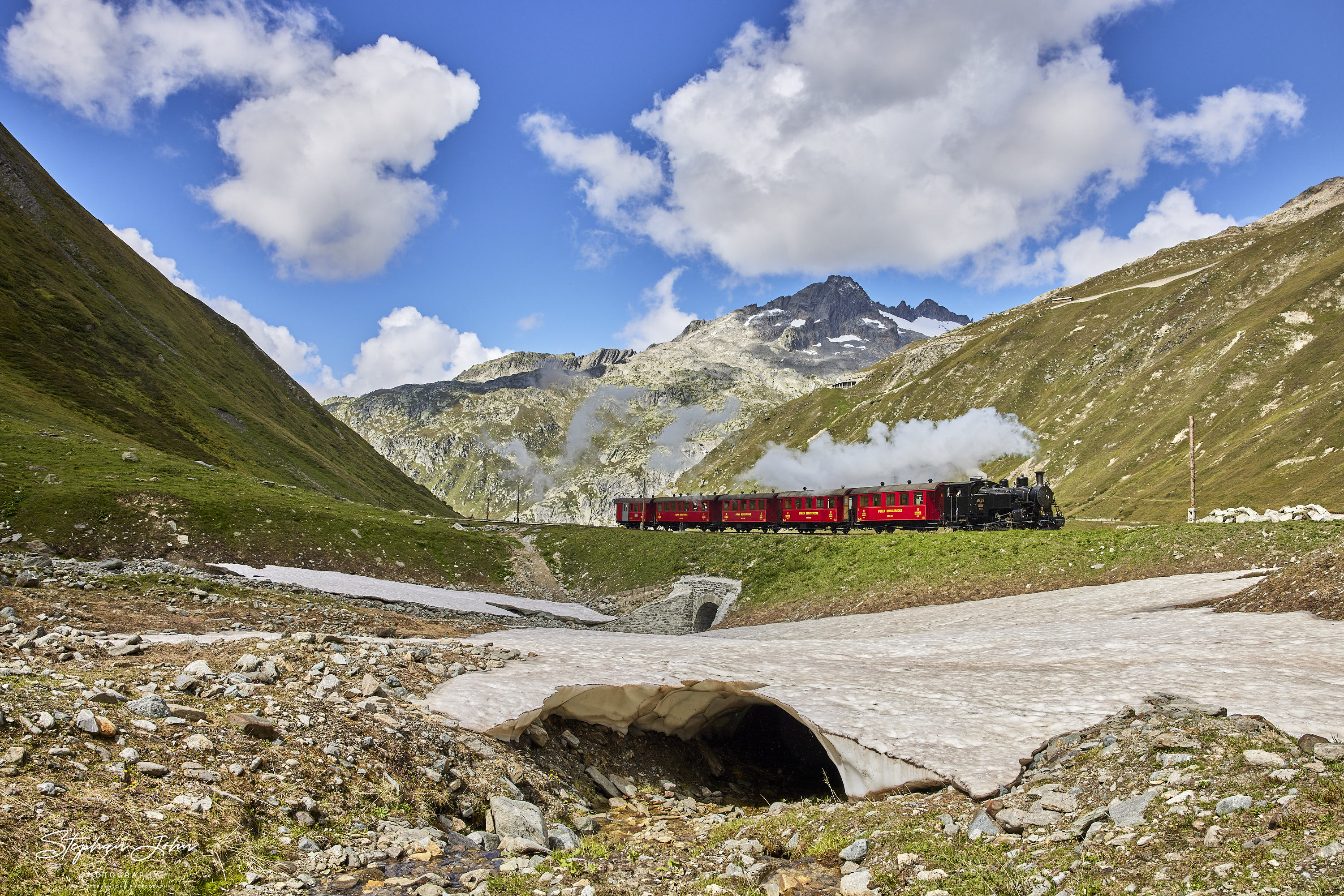 Lok 9 der DFB mit einem Personenzug fährt aus dem  Bahnhof Muttbach-Belvedere (2.120 m ü. M.) in Richtung Realp aus und verschwindet gleich im Furka-Scheiteltunnel.