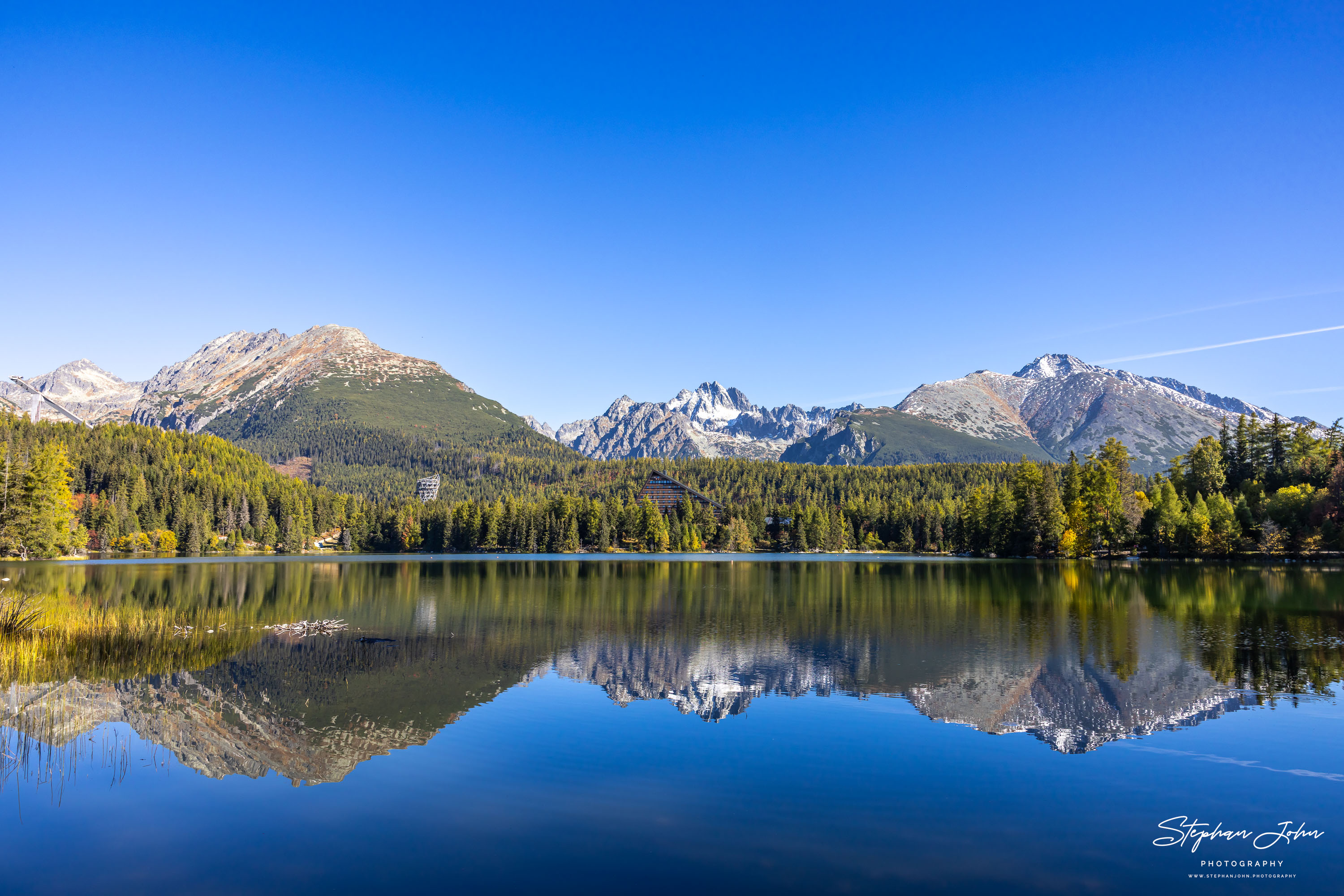 Weite Bergkette und goldgelbe Uferbäume spiegeln sich klar im blauen Štrbské pleso unter wolkenlosem Himmel.