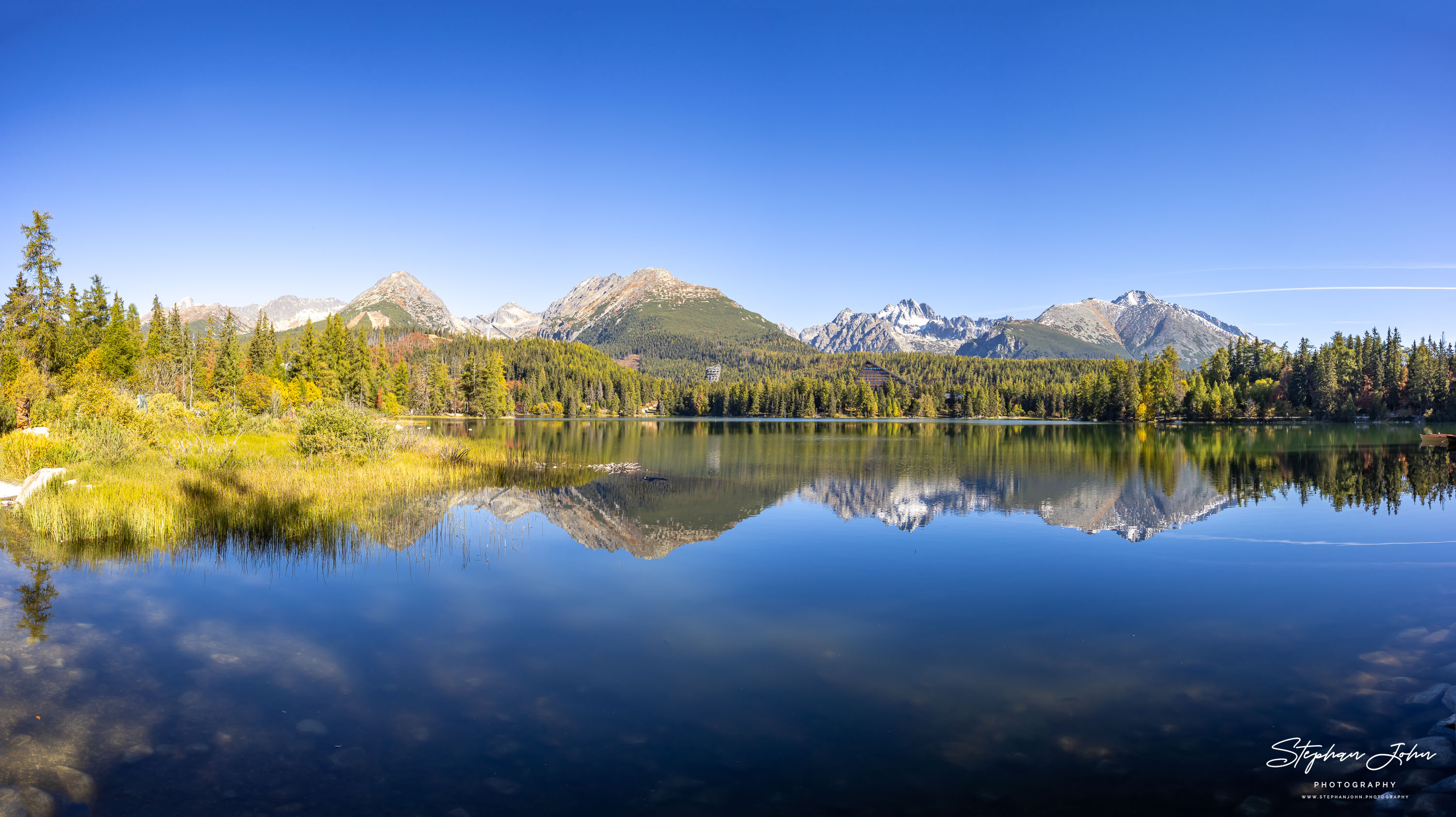 Herbstpanorama des Štrbské pleso mit goldgelbem Ufer und einer nahezu vollständigen Spiegelung von Wald und Bergkette.
