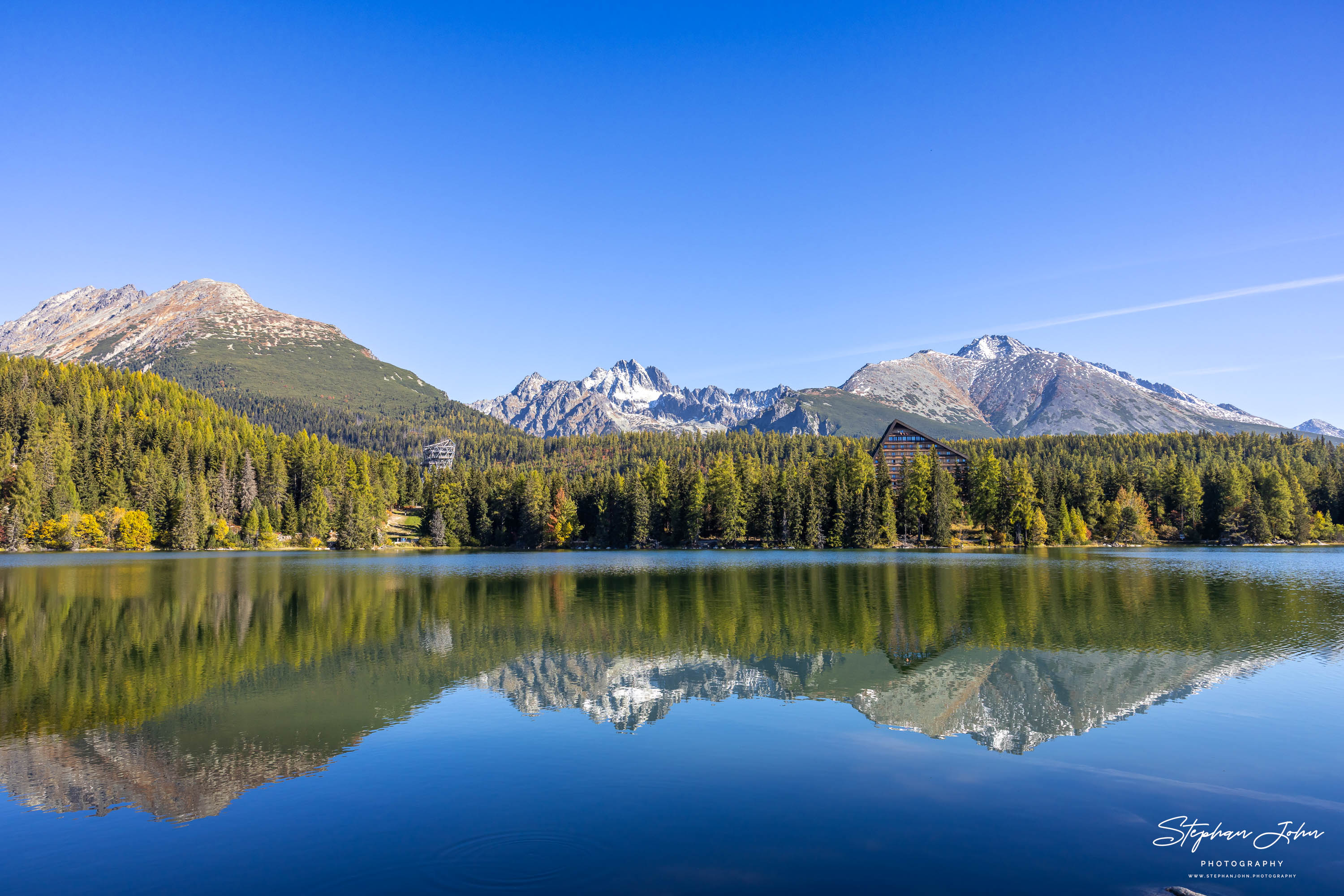 Hotel Patria zwischen Wald und Tatragipfeln am Štrbské pleso mit klarer Bergspiegelung und gelben Uferbäumen.