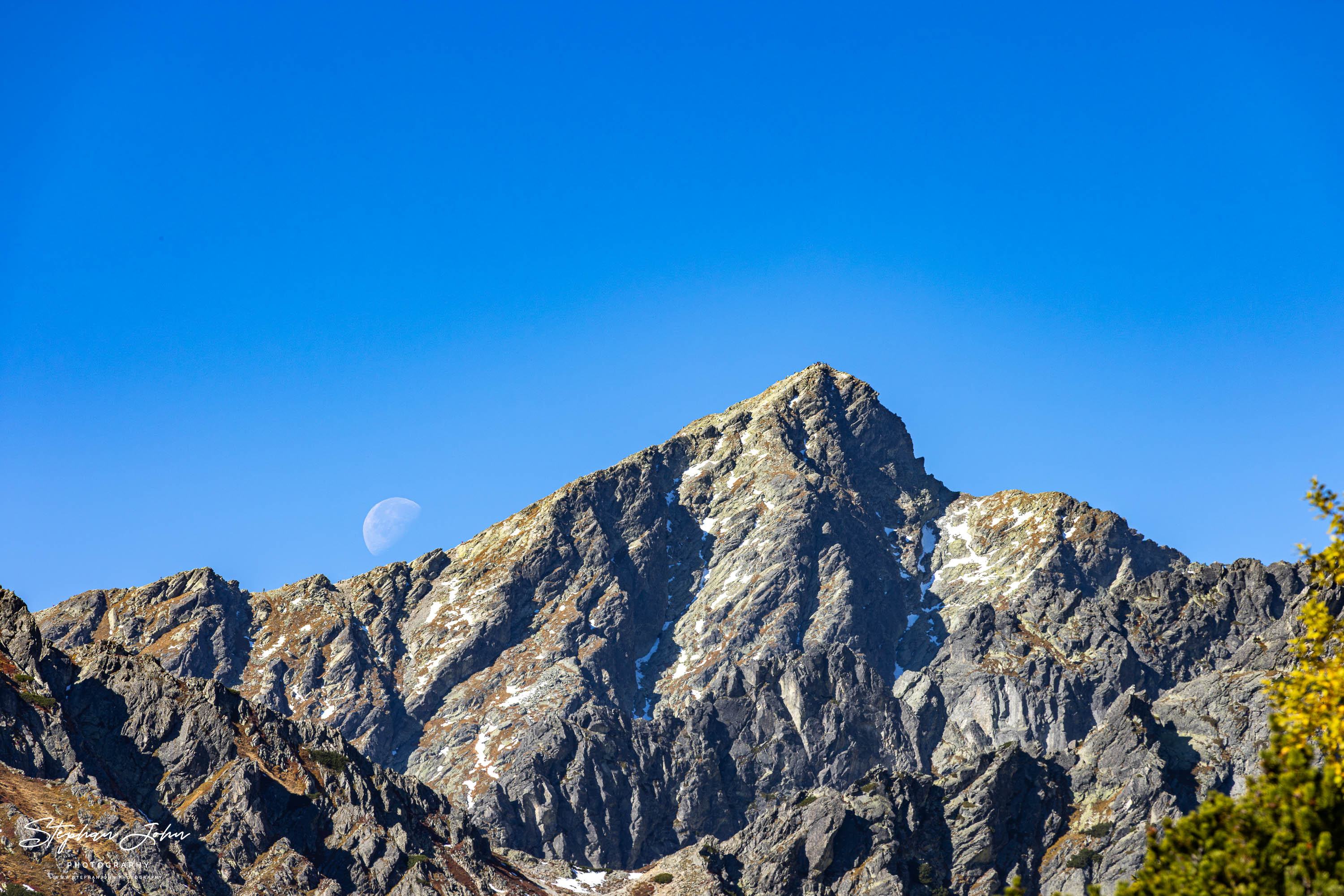 Blasser Mond links neben dem felsigen Gipfel des Kriváň, mit tiefen Rinnen und kleinen Schneeresten unter blauem Himmel.