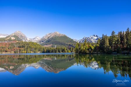 Spiegelglatter Štrbské pleso im Oktober mit klaren Spiegelbildern der Berge und des teilweise herbstlich gefärbten Waldes.