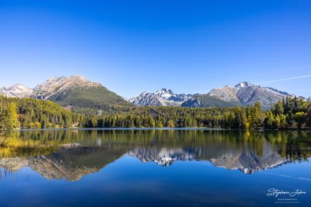 Weite Bergkette und goldgelbe Uferbäume spiegeln sich klar im blauen Štrbské pleso unter wolkenlosem Himmel.