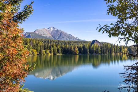 Orangefarbenes Laub und grüne Zweige rahmen den Štrbské pleso mit Hotel Patria und einer ruhigen Bergspiegelung ein.