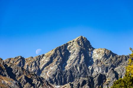 Blasser Mond links neben dem felsigen Gipfel des Kriváň, mit tiefen Rinnen und kleinen Schneeresten unter blauem Himmel.